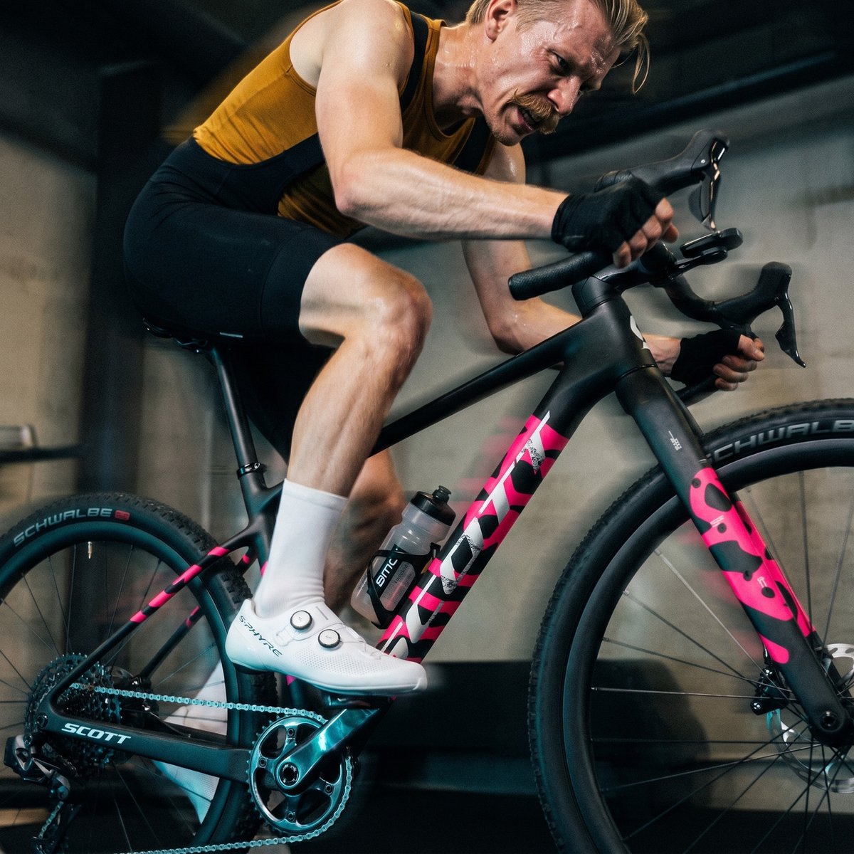 A rider in a mustard vest on a gravel bike, knobbly tyres still fitted, set up on a trainer in a bare concrete garage.